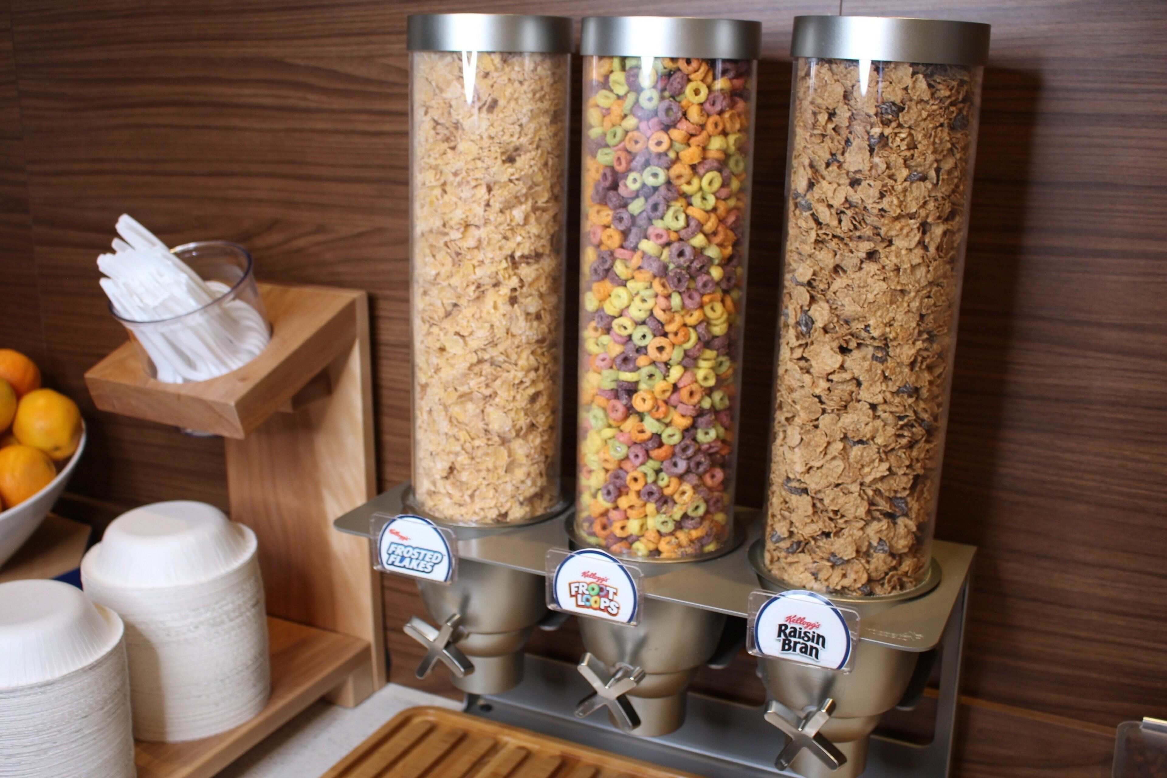 Tres dispensadores cilíndricos con etiquetas para distintos tipos de cereal, un cuenco de naranjas y un rodillo de toallas de papel.