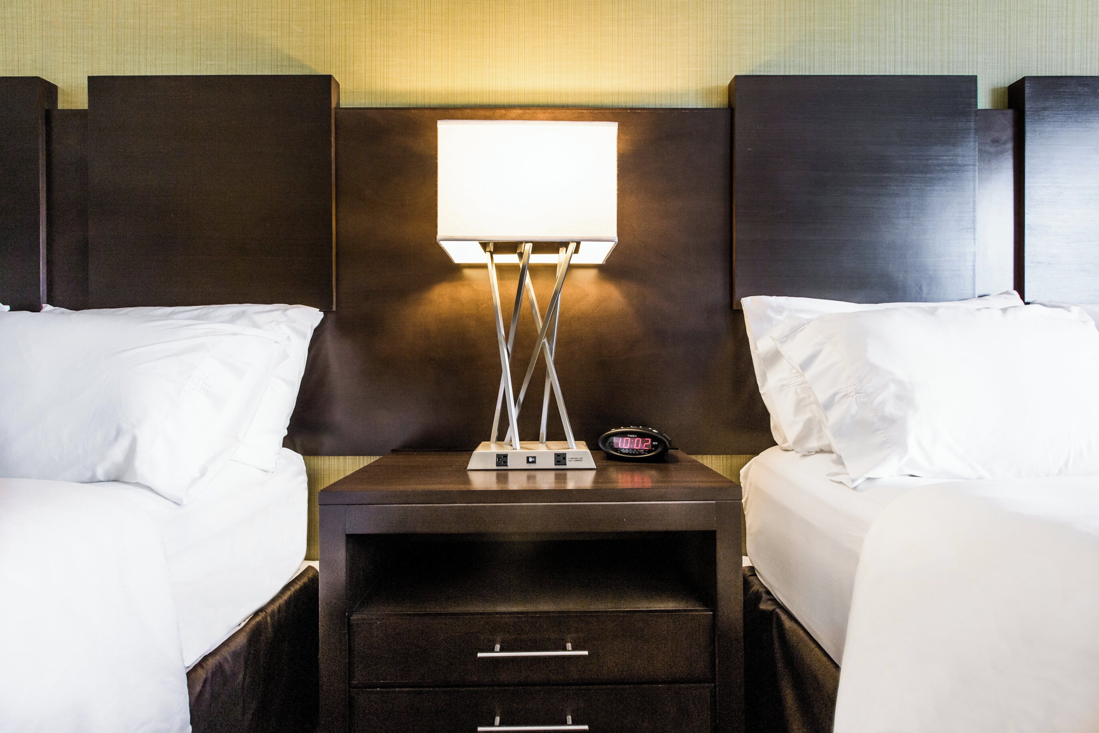 Et hotelværelse med to senge, et natbord med skuffer, en lampe og et digitalt ur.