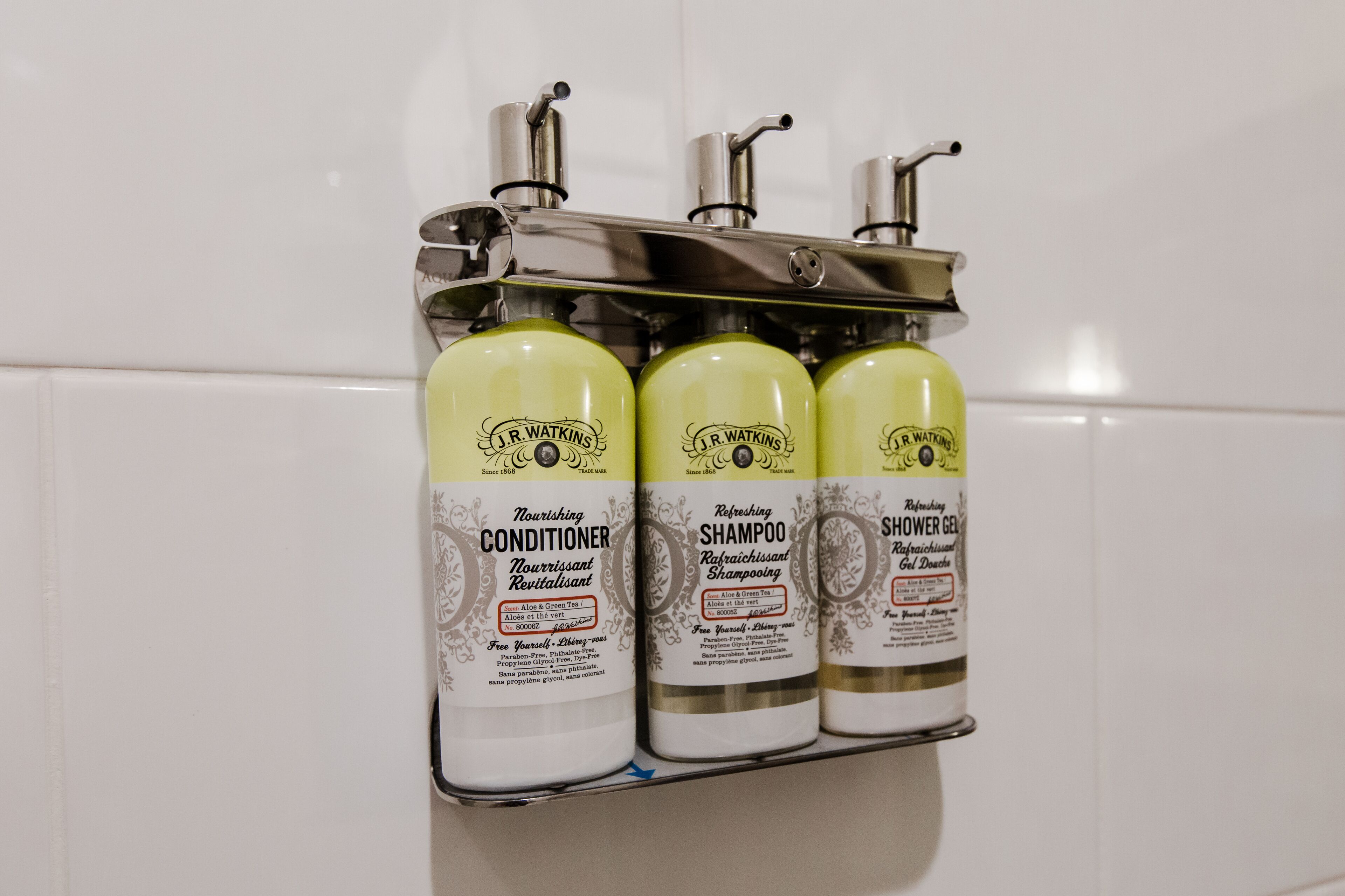 Tre flaconi di Balsamo Nutriente, Shampoo e Gel Doccia J.R. Watkins su un dispenser metallico in un bagno.