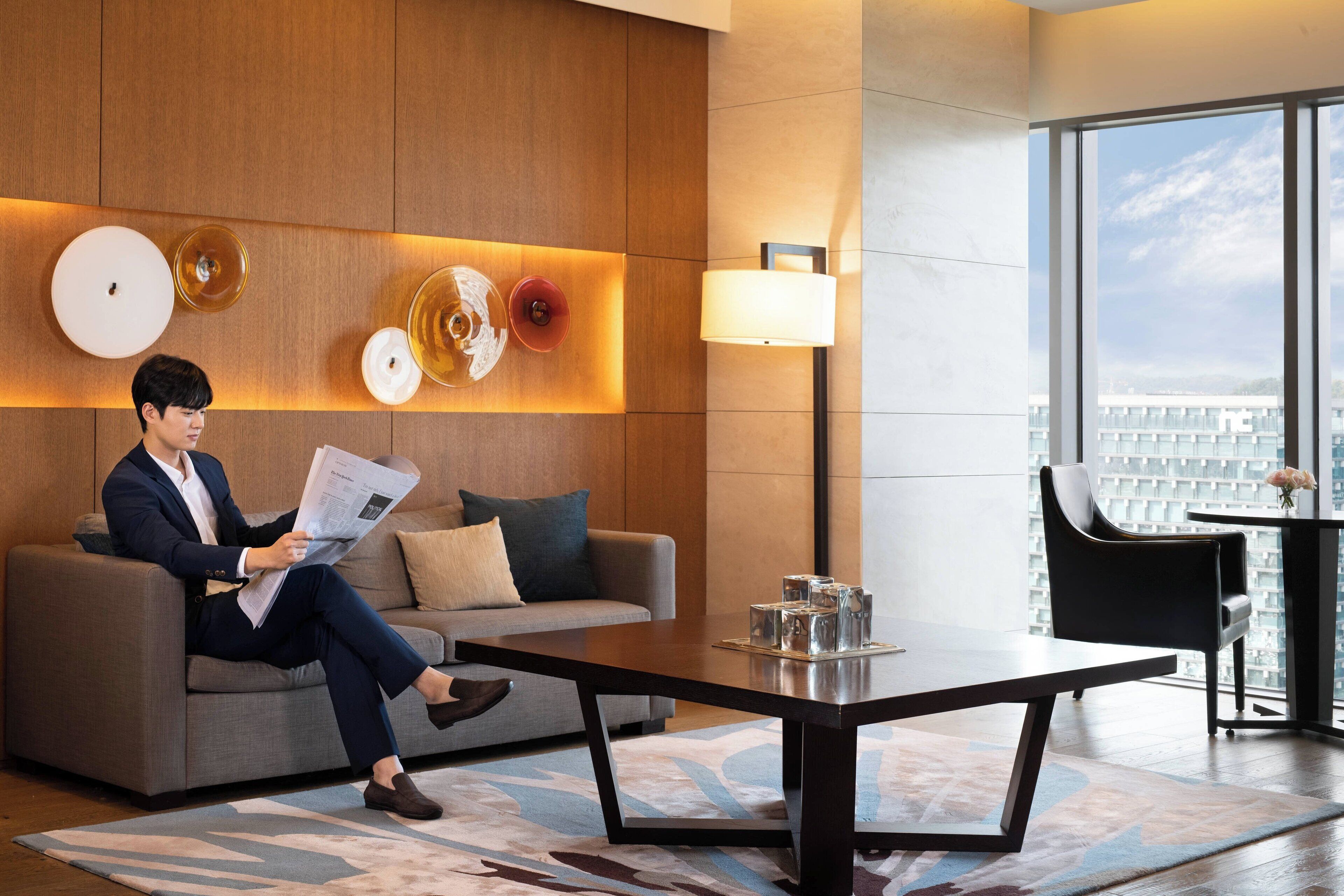 A modern hotel room with a sofa, a coffee table, a lamp, and a view of the city through large windows.