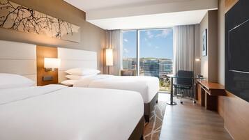Habitación de hotel con dos camas, un escritorio, una silla, un televisor y un ventanal con vistas a la ciudad.