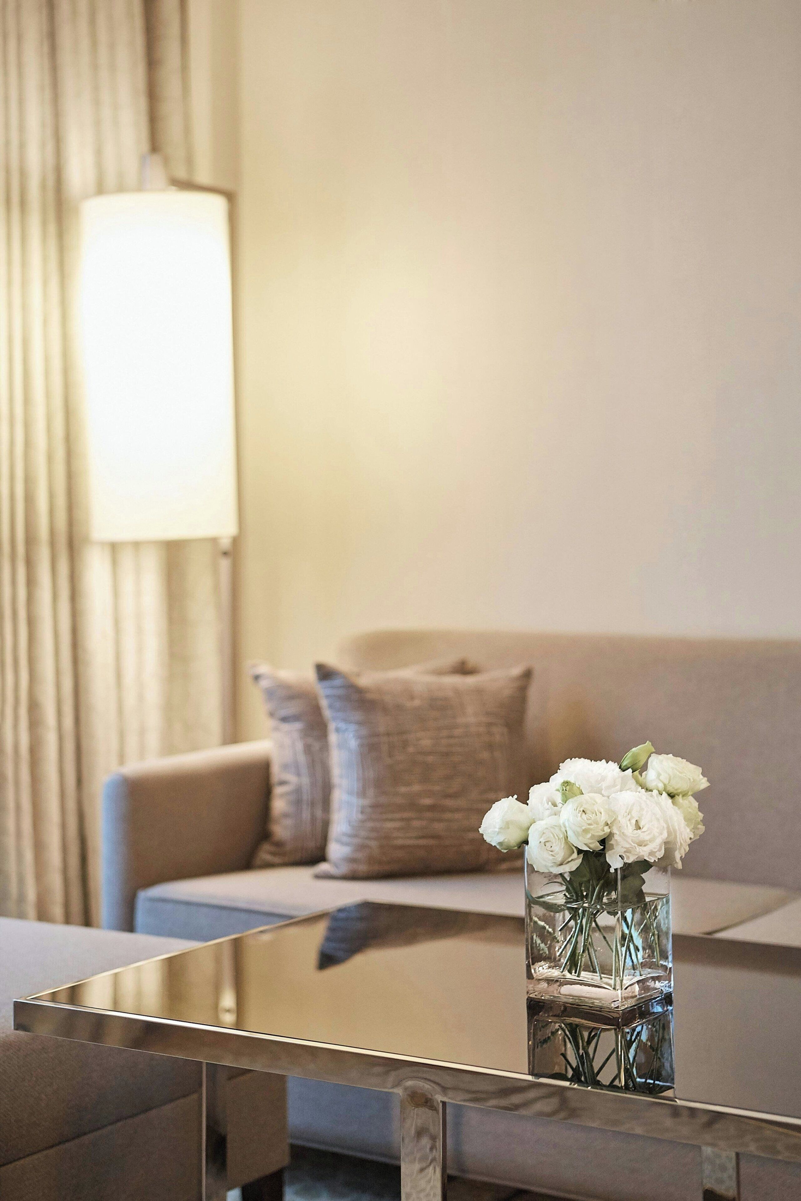 Un salon moderne avec une table basse en verre, un canapé beige agrémenté de coussins et un vase contenant des fleurs blanches.