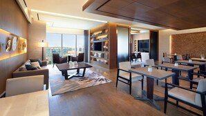 A modern hotel lobby with a sofa, dining tables, chairs, a bookshelf, and a city view.