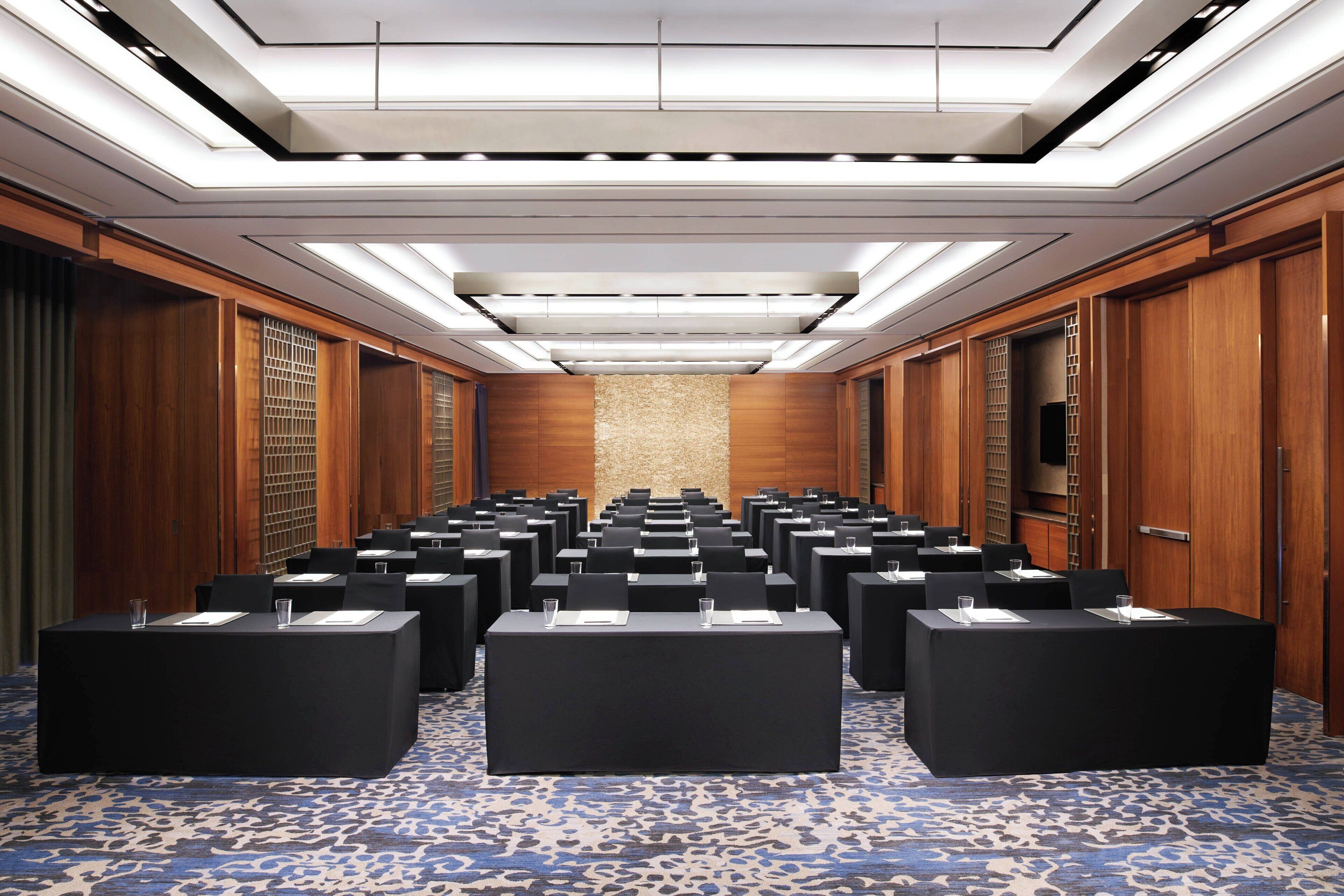 A conference room with rows of tables and chairs, wooden paneling, and a patterned carpet.