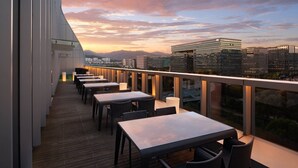 A rooftop terrace with tables and chairs, overlooking a cityscape and mountains during sunset.