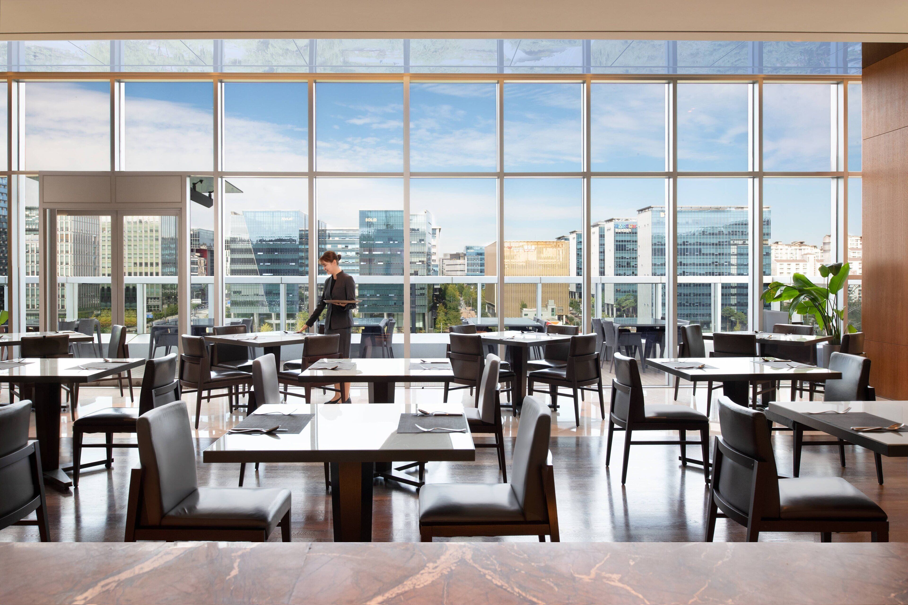 A modern restaurant with large windows offering a city view, neatly arranged tables and chairs, and a clean, contemporary interior design.