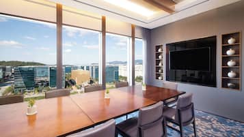 Un salón de conferencias con una gran mesa, sillas, una pantalla de televisión y vistas a la ciudad a través de amplias ventanas.