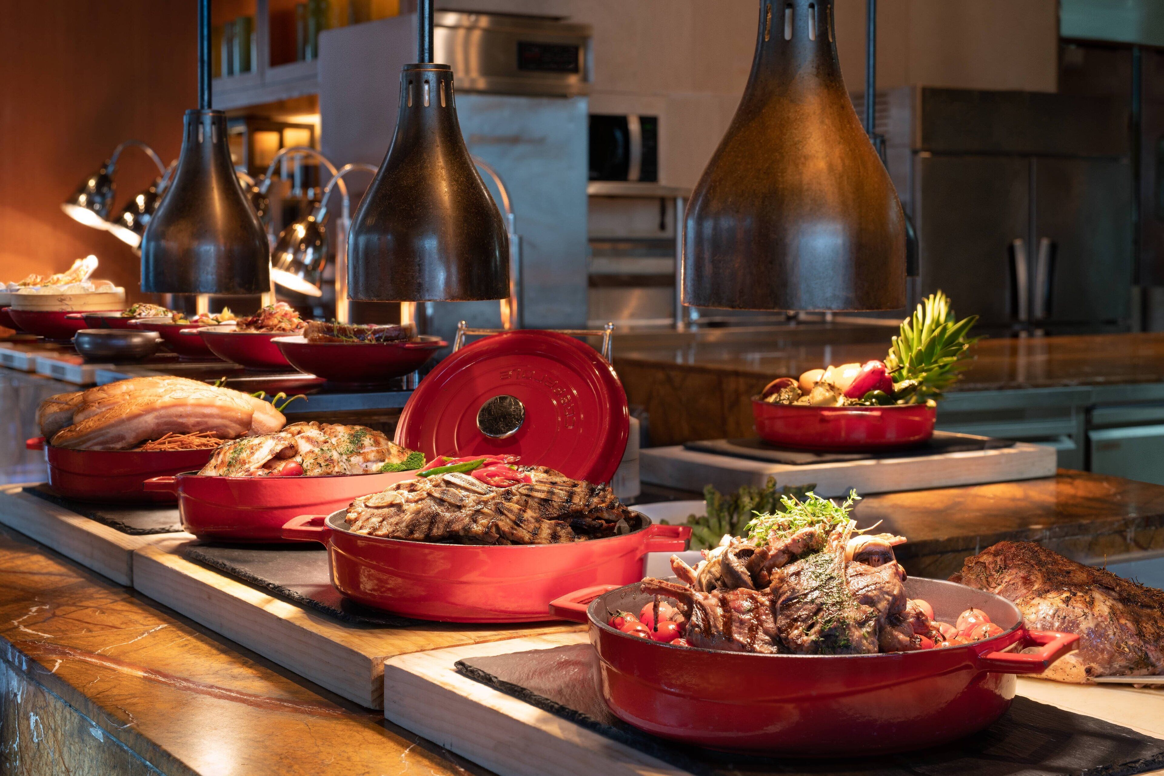 Ein Buffet mit verschiedenen Gerichten in roten Töpfen, darunter gegrilltes Fleisch und Gemüse, auf einem Holztisch in einer modernen Küche.
