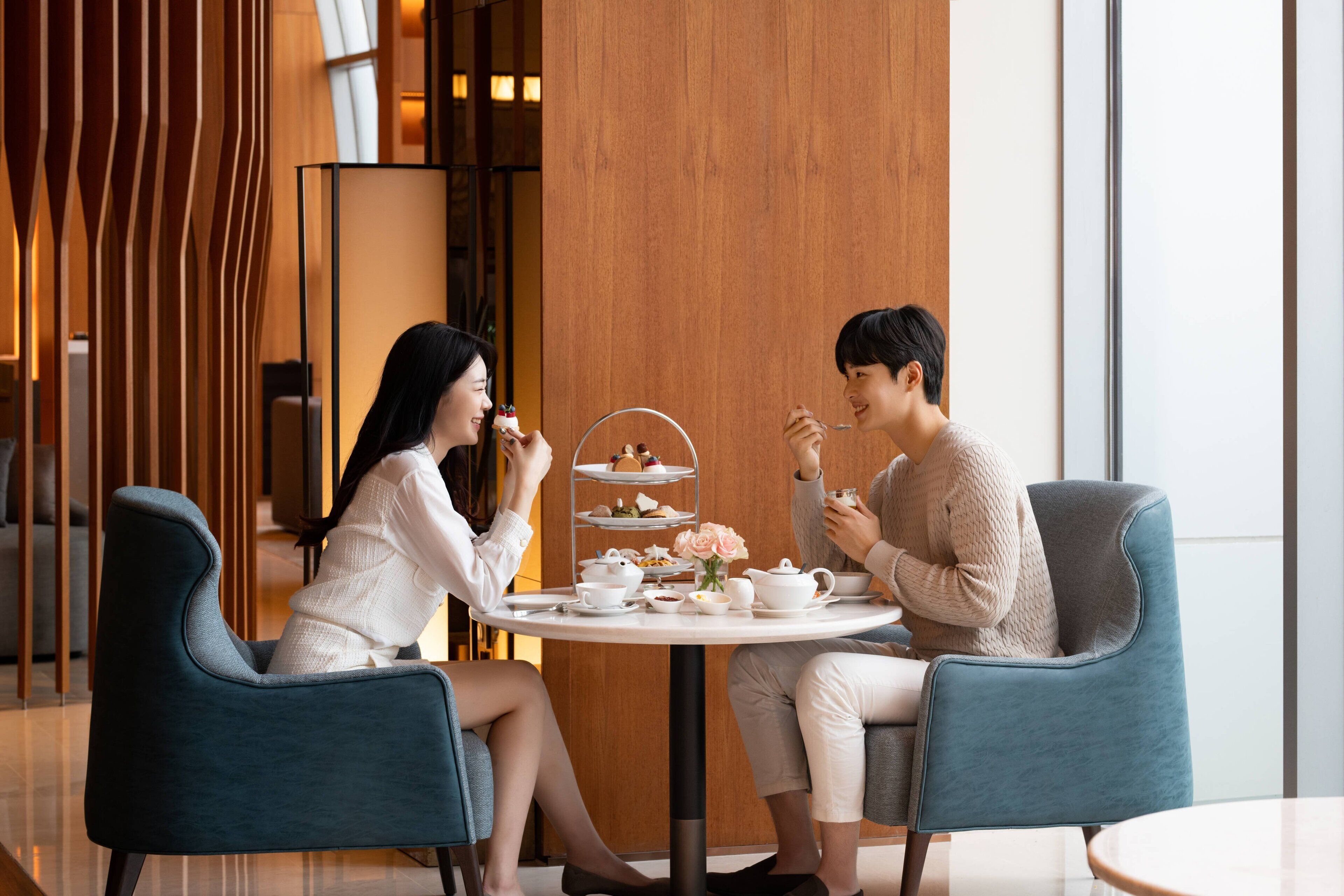 A modern interior with two people seated at a round table with a tea set, surrounded by blue chairs, in a room with wooden paneling and large windows.