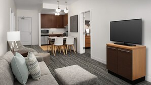 Ein modernes Wohnzimmer mit Sofa, Hocker und einem Flachbildfernseher auf einem Holzschrank. Im Hintergrund ist ein Essbereich mit Stühlen und Tisch zu sehen.