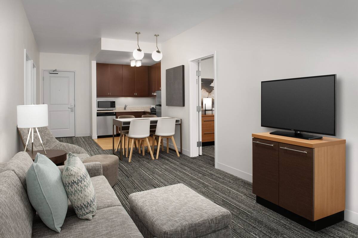 A modern living room with a sofa, ottoman, and a flat-screen TV on a wooden cabinet. A dining area with chairs and a table is visible in the background.