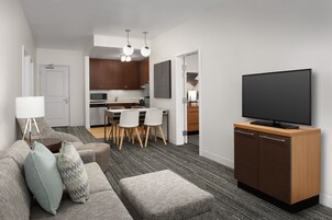 Una sala moderna con un sofá, un reposapiés y un televisor de pantalla plana sobre un mueble de madera. Se ve un área de comedor con sillas y una mesa al fondo.