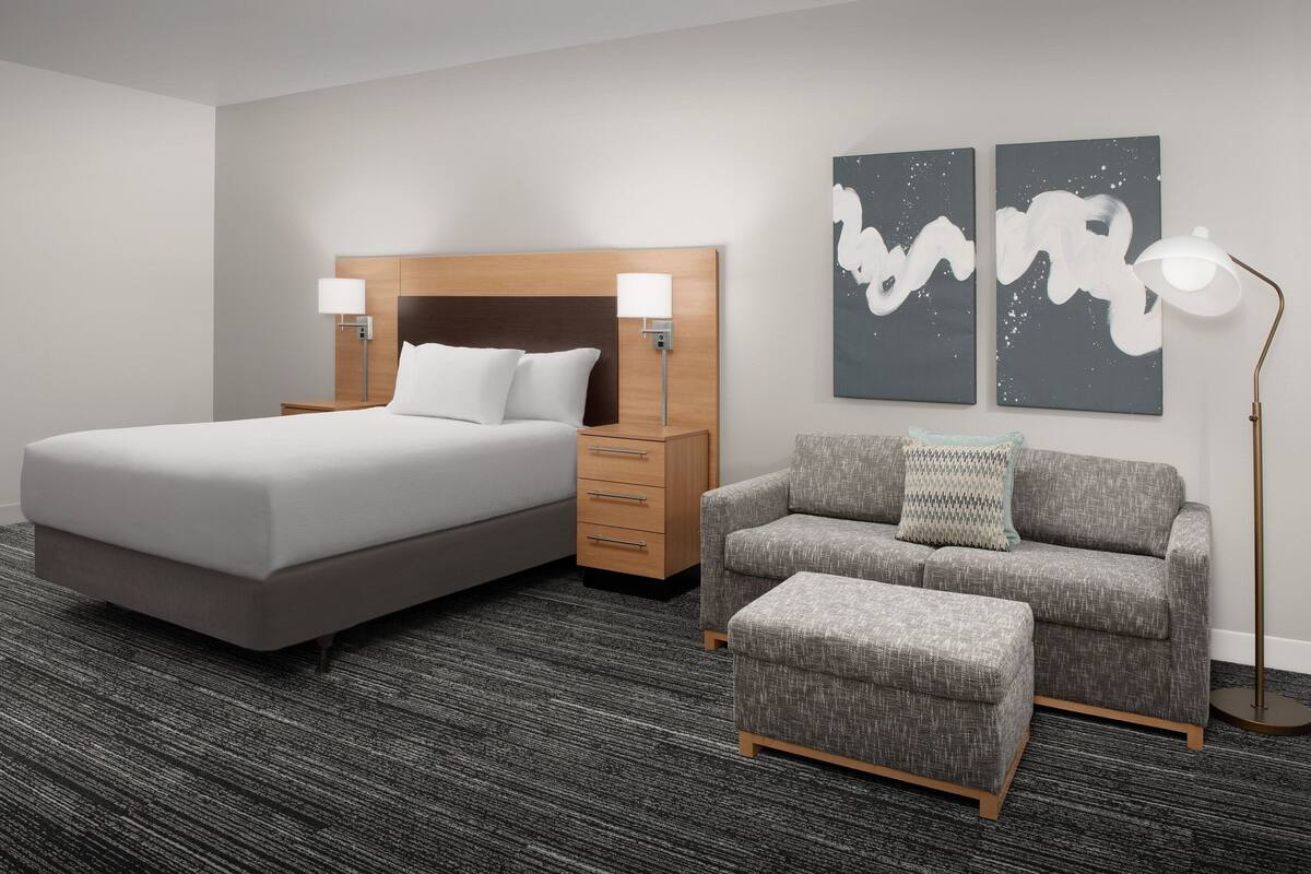 A hotel room with a large bed, a sofa, a small ottoman, a nightstand, and two wall-mounted lamps.