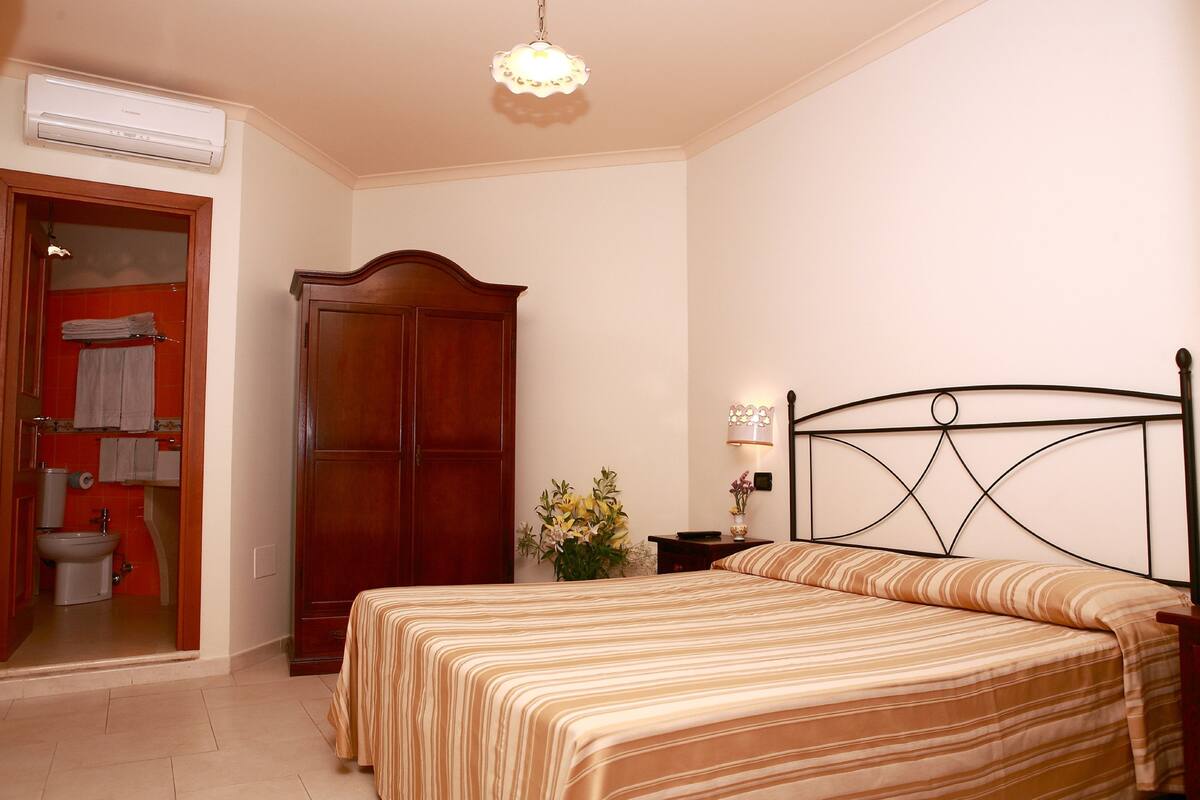 A bedroom with a bed, a wooden wardrobe, a nightstand, a lamp, and a bathroom visible through an open door.