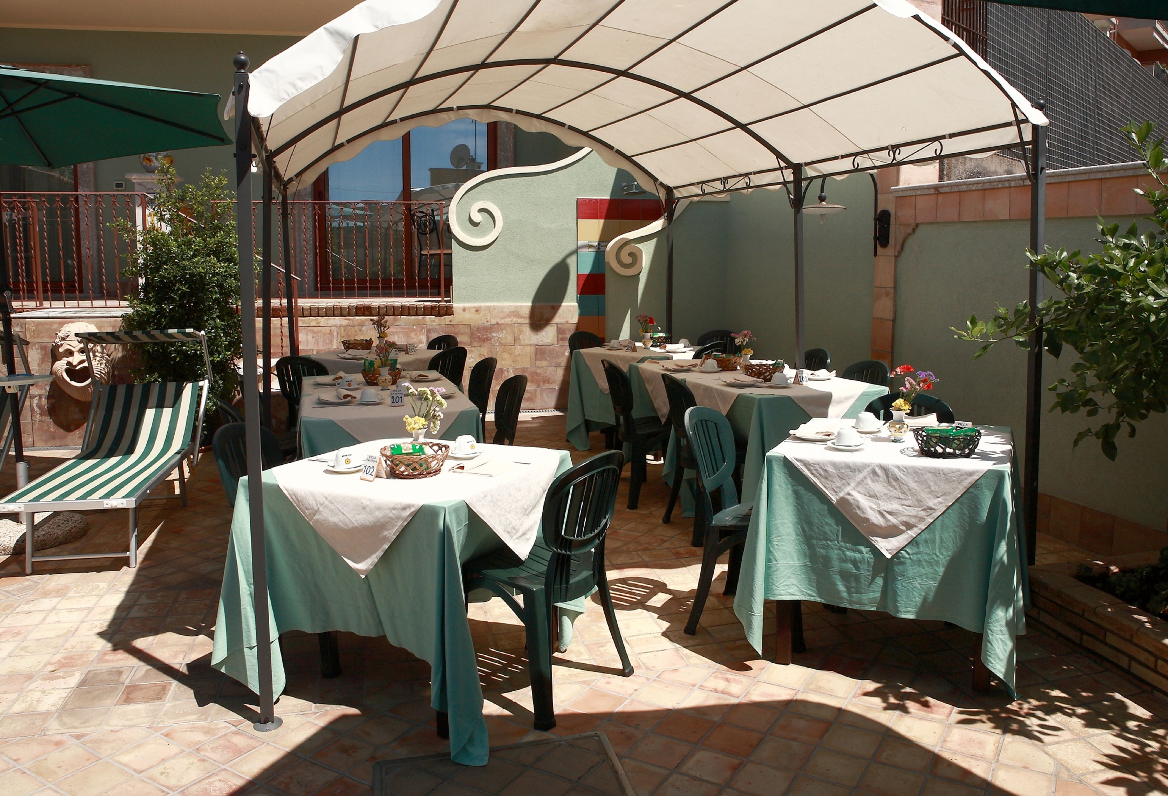 Un área de comedor al aire libre con mesas cubiertas de manteles blancos, sillas verdes y una gran sombrilla que da sombra.