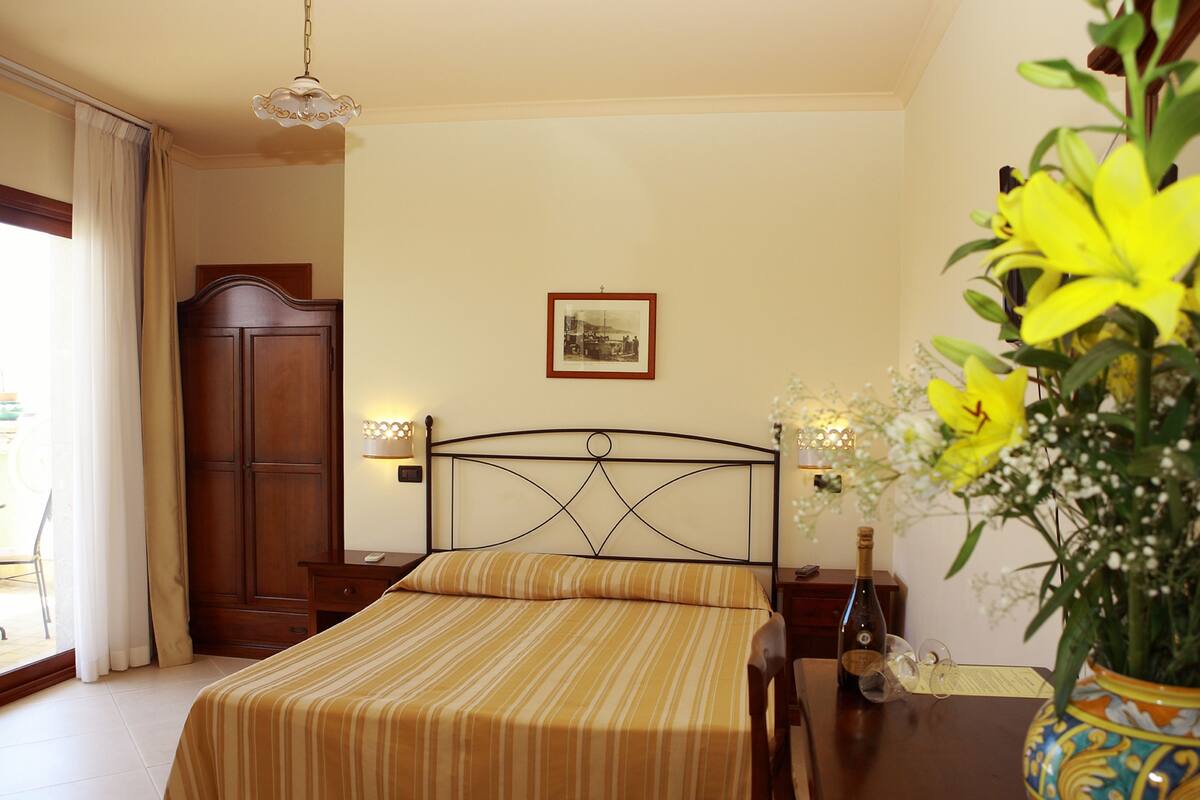 A hotel room with a bed, a wooden wardrobe, a nightstand with a lamp, a framed picture on the wall, and a vase with flowers on a table.