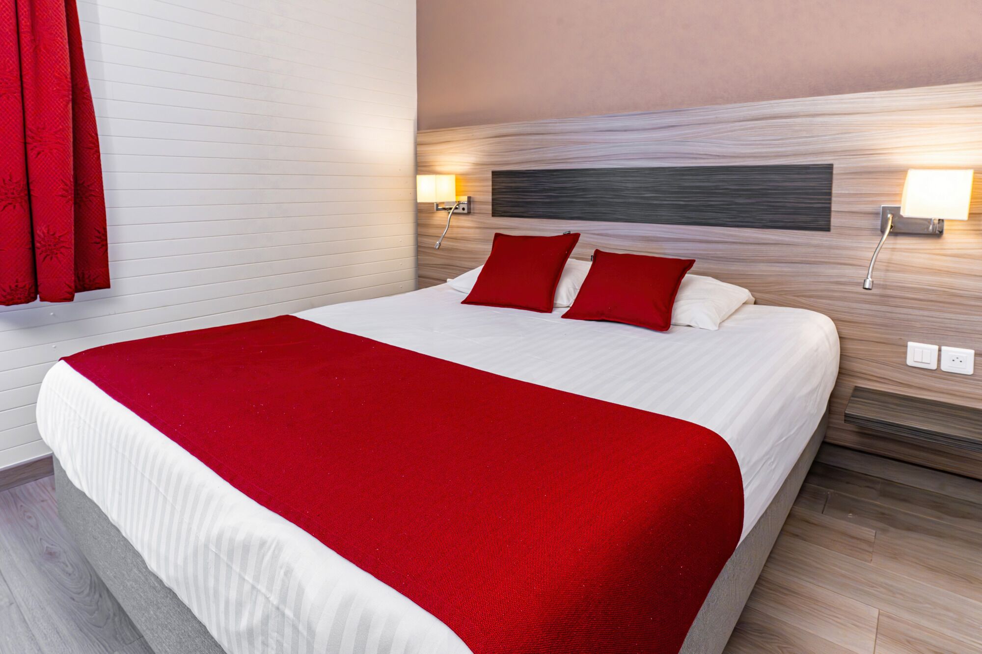 Quarto de hotel com uma cama grande, roupa de cama vermelha, duas luminárias de cabeceira e uma cabeceira de madeira.