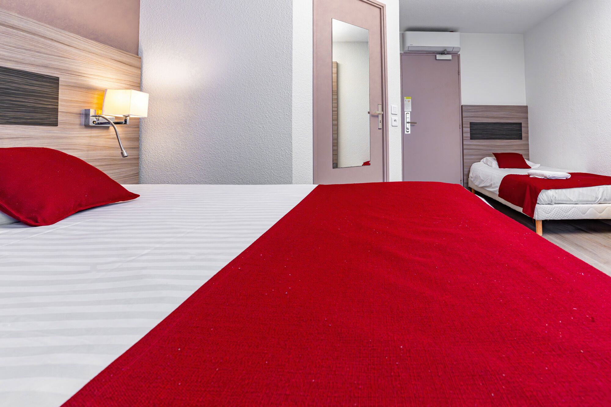 A hotel room with two beds, a red throw blanket, a nightstand with a lamp, and a mirror.