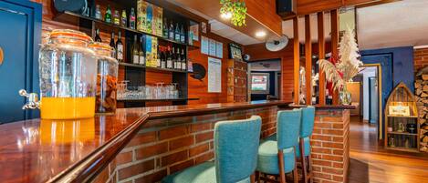 A bar area with wooden walls, a brick counter, and blue bar stools. There are shelves with bottles and glasses, and a hanging plant.