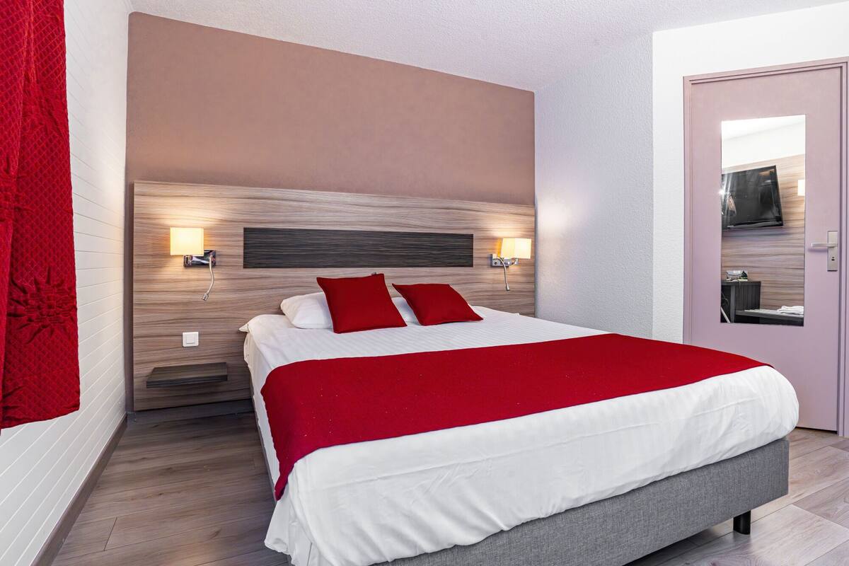 A modern hotel room with a large bed, red and white bedding, two wall-mounted lights, and a wooden headboard.