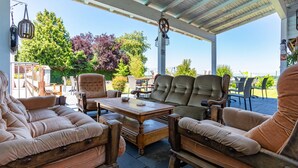 Un patio exterior con mobiliario de madera, incluyendo sofás y sillas, bajo una zona cubierta con una rueda decorativa y faroles colgantes.