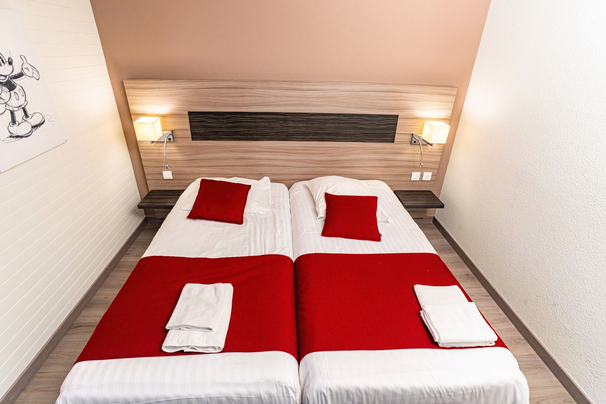 A double bed with two red pillows and white linens, a wooden headboard, bedside lamps, and a red carpet.