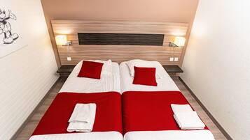 A double bed with two red pillows and white linens, a wooden headboard, bedside lamps, and a red carpet.
