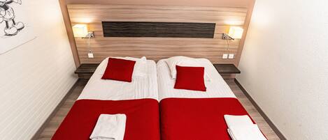 A double bed with two red pillows and white linens, a wooden headboard, bedside lamps, and a red carpet.