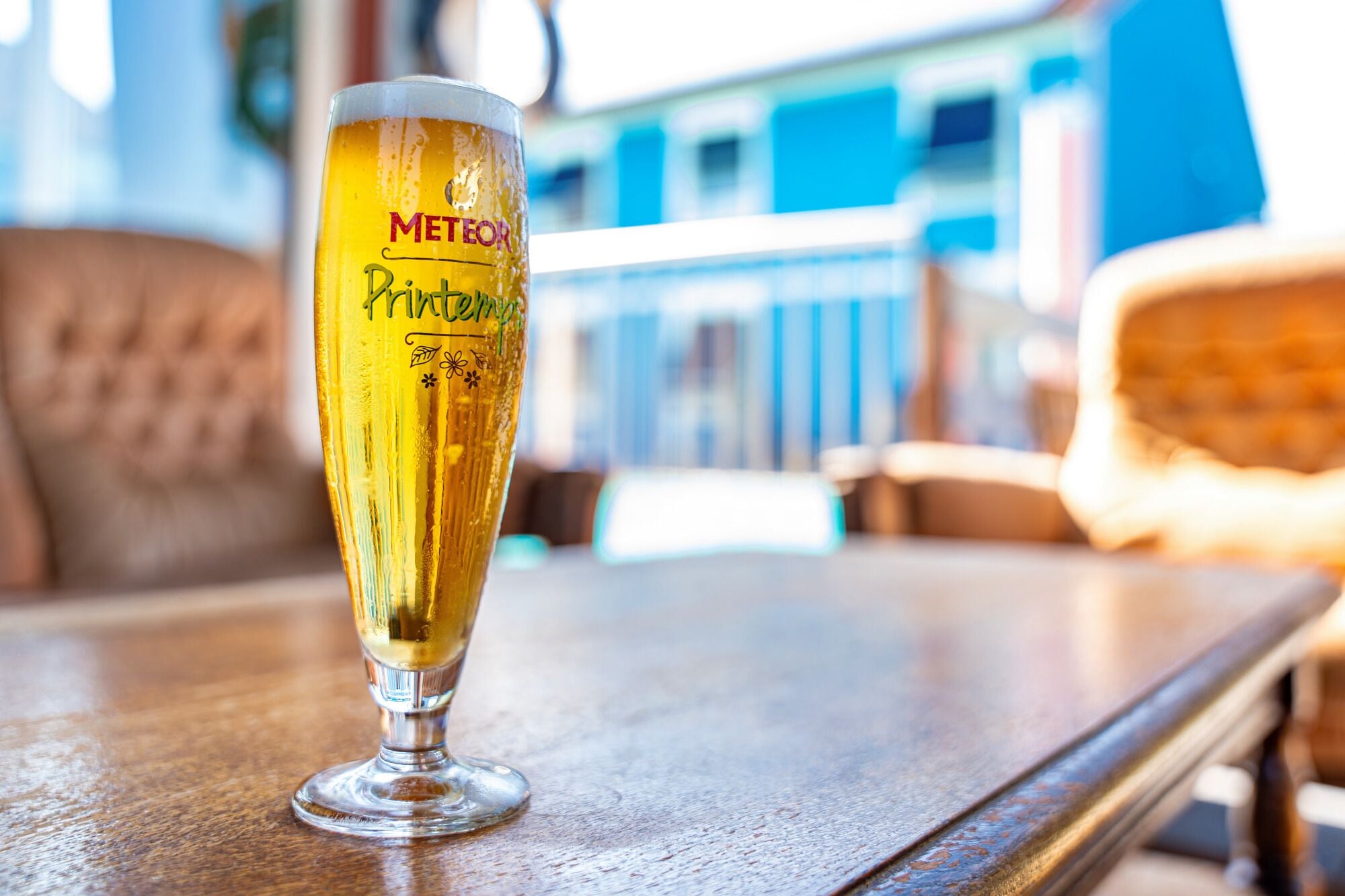 A glass of beer with "METEO" and "Pintemps" on it, placed on a wooden table in a room with a blurred background.