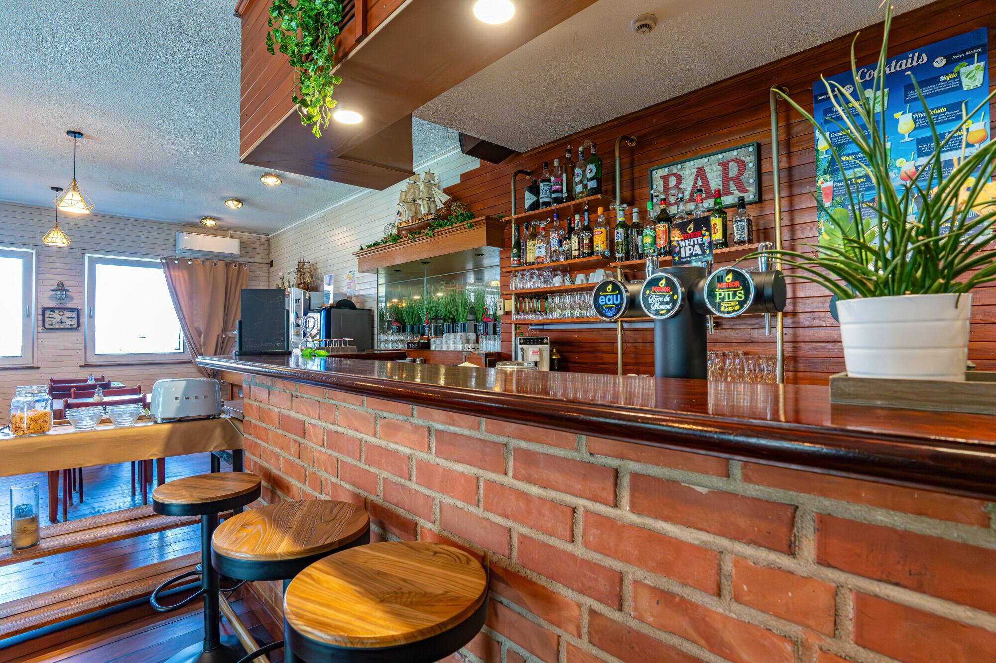 Zona de bar con pared de ladrillo, taburetes de madera, mostrador con caja registradora y un tablero de menú.