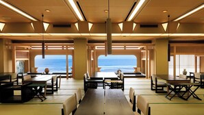 Un comedor amplio con mesas y sillas, ventanas grandes con vistas al océano y un techo alto con iluminación lineal.