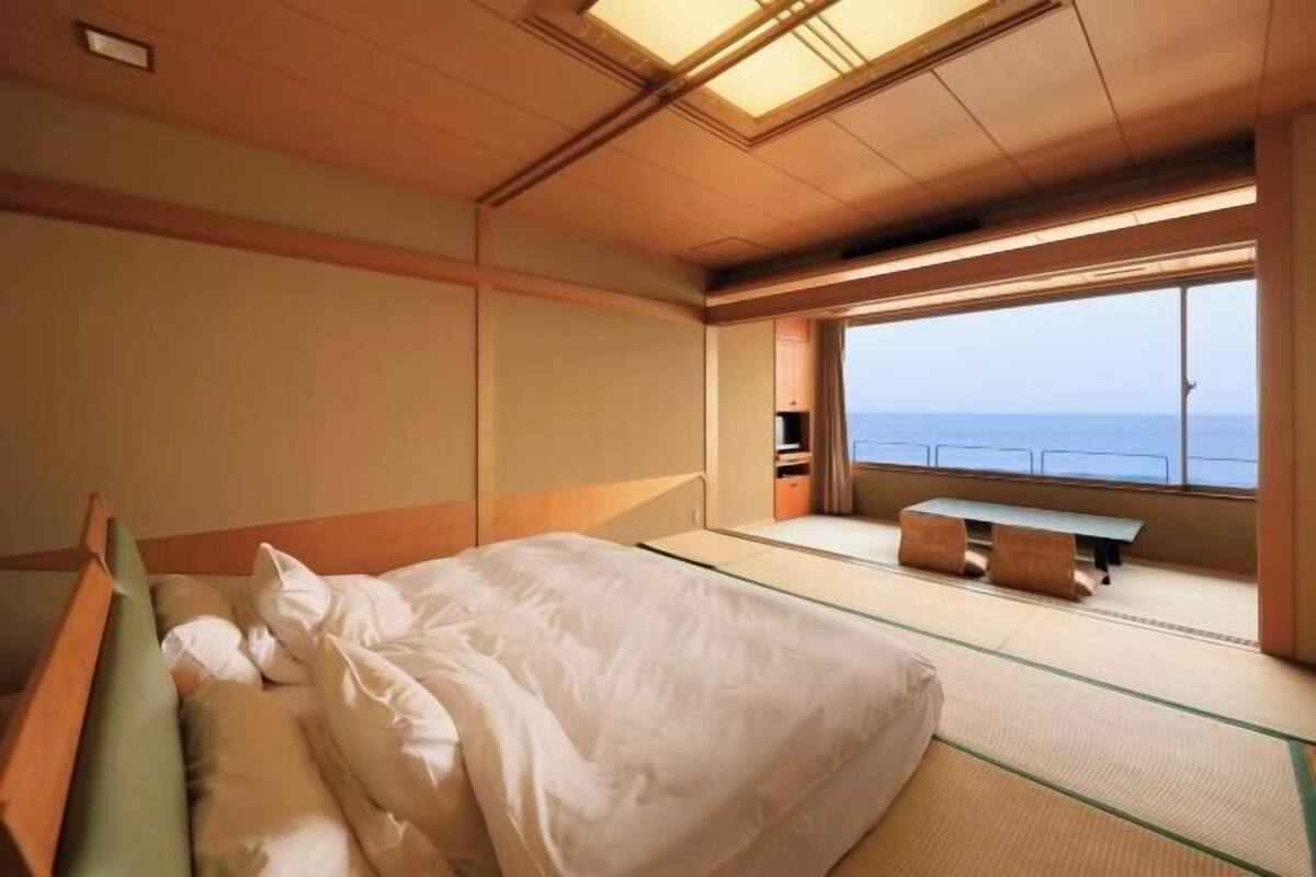 A room with a bed, a small table, and a window with a view of the sea.