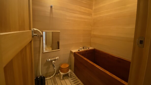 A wooden-paneled bathroom with a wooden bathtub, a small wooden stool, a mirror, and a wall-mounted heater. - Wakamatsu Hot Spring Resort (Hakodate)