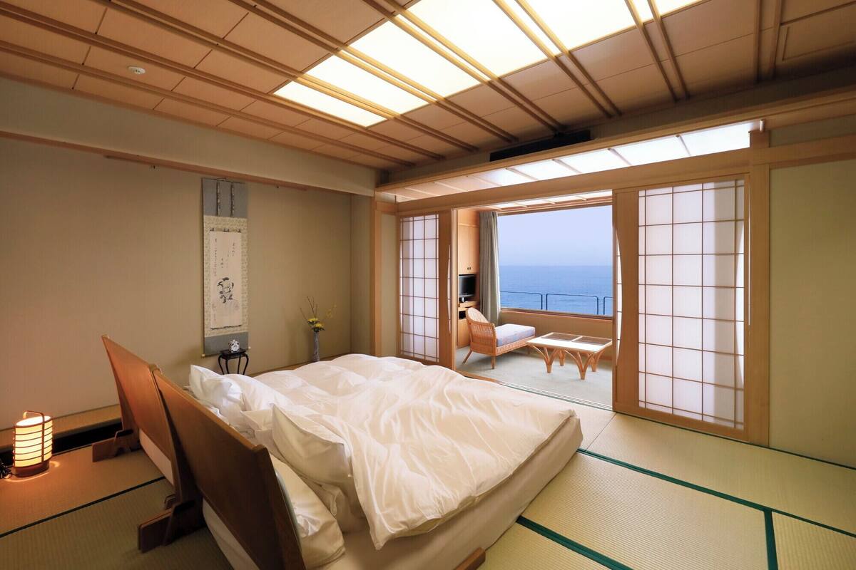 A traditional Japanese-style room with tatami flooring, a bed, a TV, and a view of the sea.