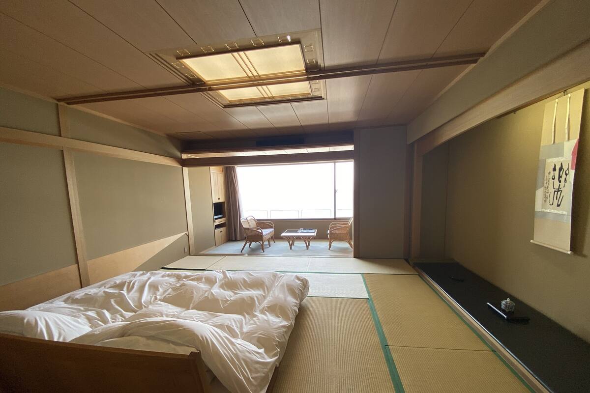 A traditional Japanese room with tatami flooring, a futon bed, a sliding door leading to a balcony, and a view of the outside.