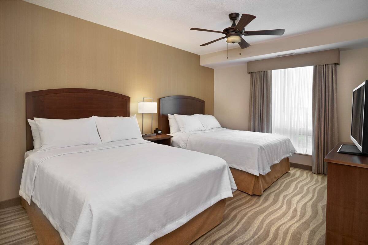 A hotel room with two beds, a ceiling fan, a TV, and a window with curtains.