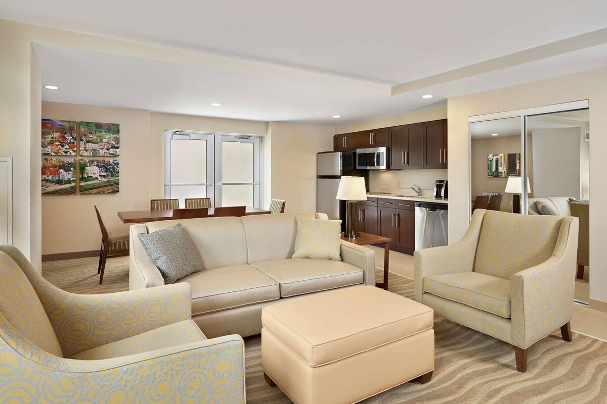 A modern living room with a sofa, armchairs, and a coffee table. There is a kitchen area with cabinets and appliances.