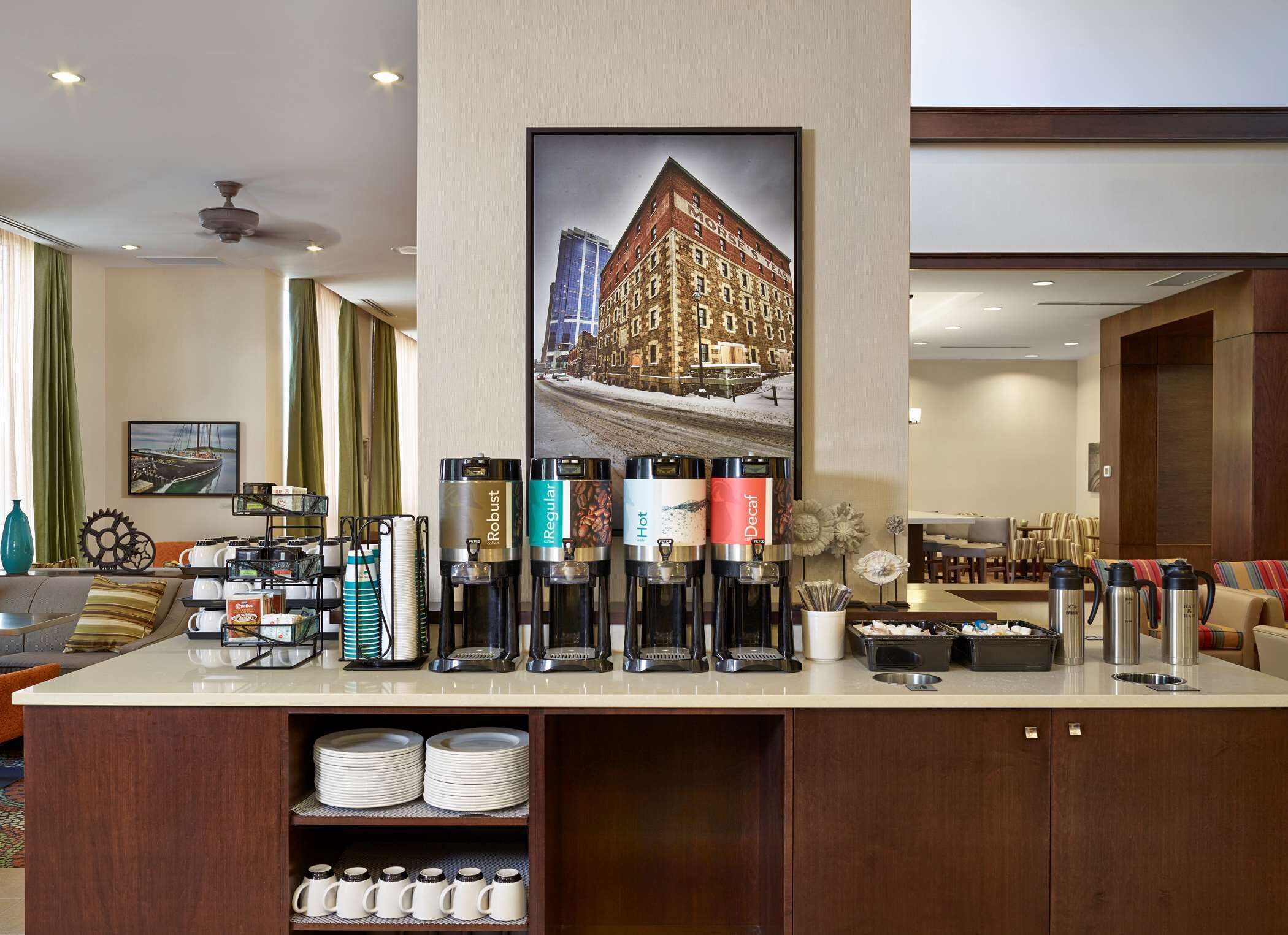 A hotel lobby with a coffee station, a large framed picture of a building, and a seating area with a sofa and chairs.