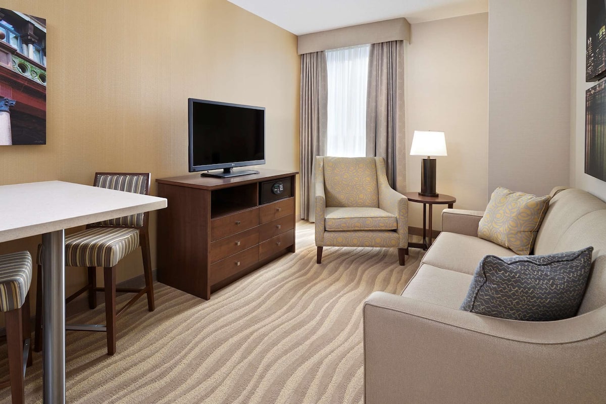 A hotel room with a flat-screen TV, a sofa, a chair, a small table with a lamp, and a dining area with chairs and a table.