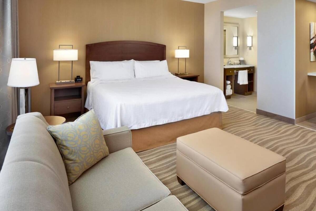 A hotel room with a bed, a sofa, an ottoman, a nightstand, a lamp, a mirror, and a bathroom visible in the background.