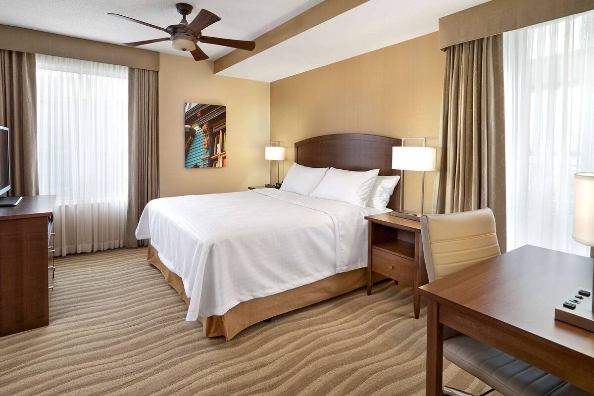 A hotel room with a bed, bedside lamps, a desk, a chair, a television, and a ceiling fan.