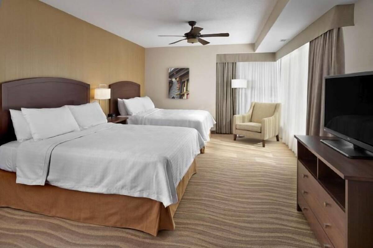 A hotel room with two beds, a TV, a ceiling fan, a chair, and a painting on the wall.