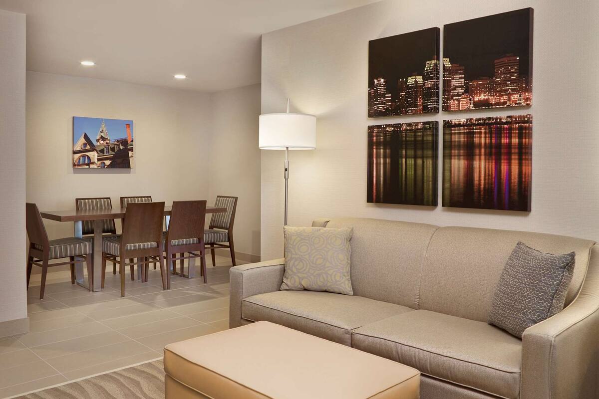 A modern living room with a sofa, ottoman, and dining set. There are three paintings on the wall, one of a cityscape at night and two abstract pieces.