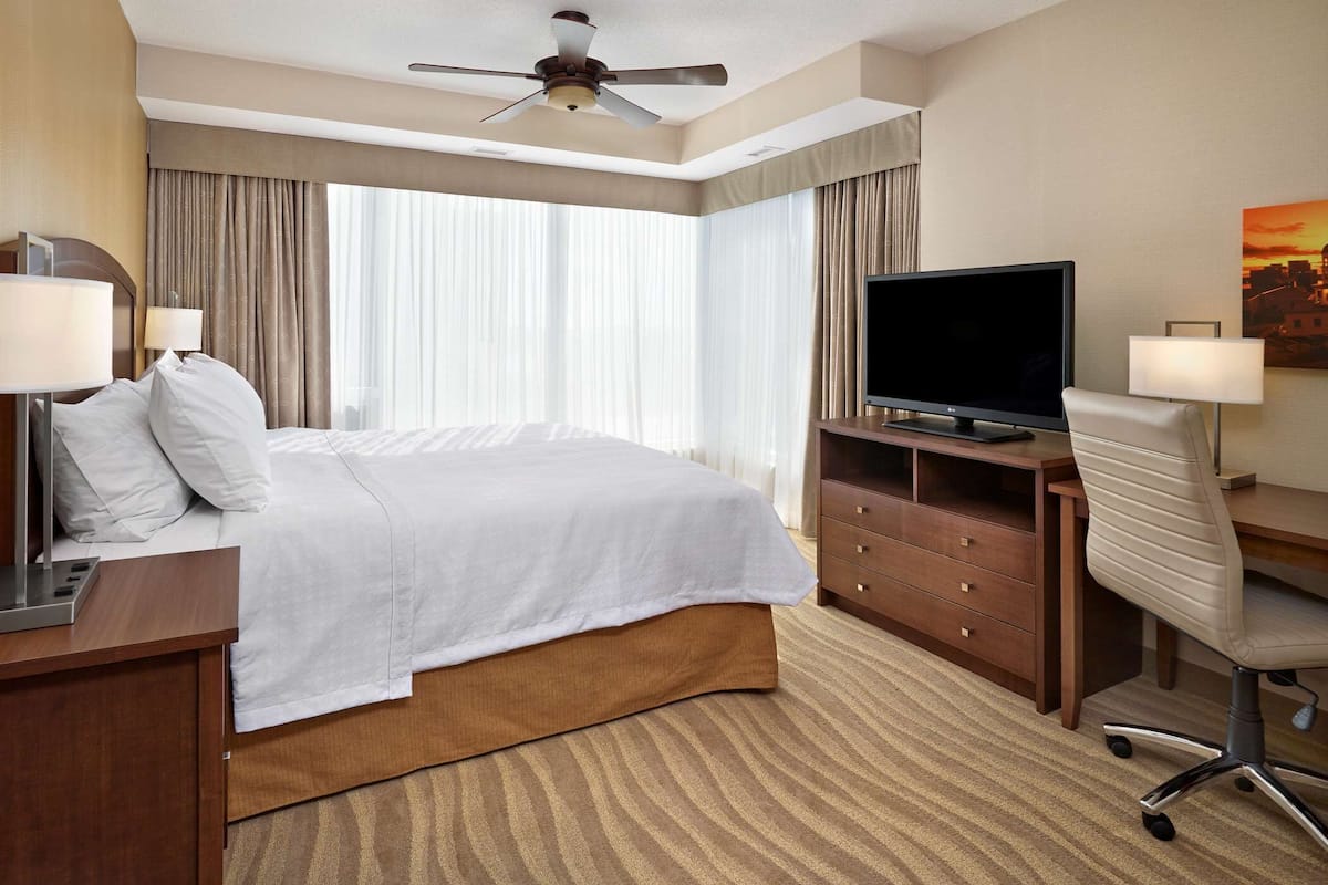 A hotel room with a bed, a desk with a television, a chair, and a ceiling fan.