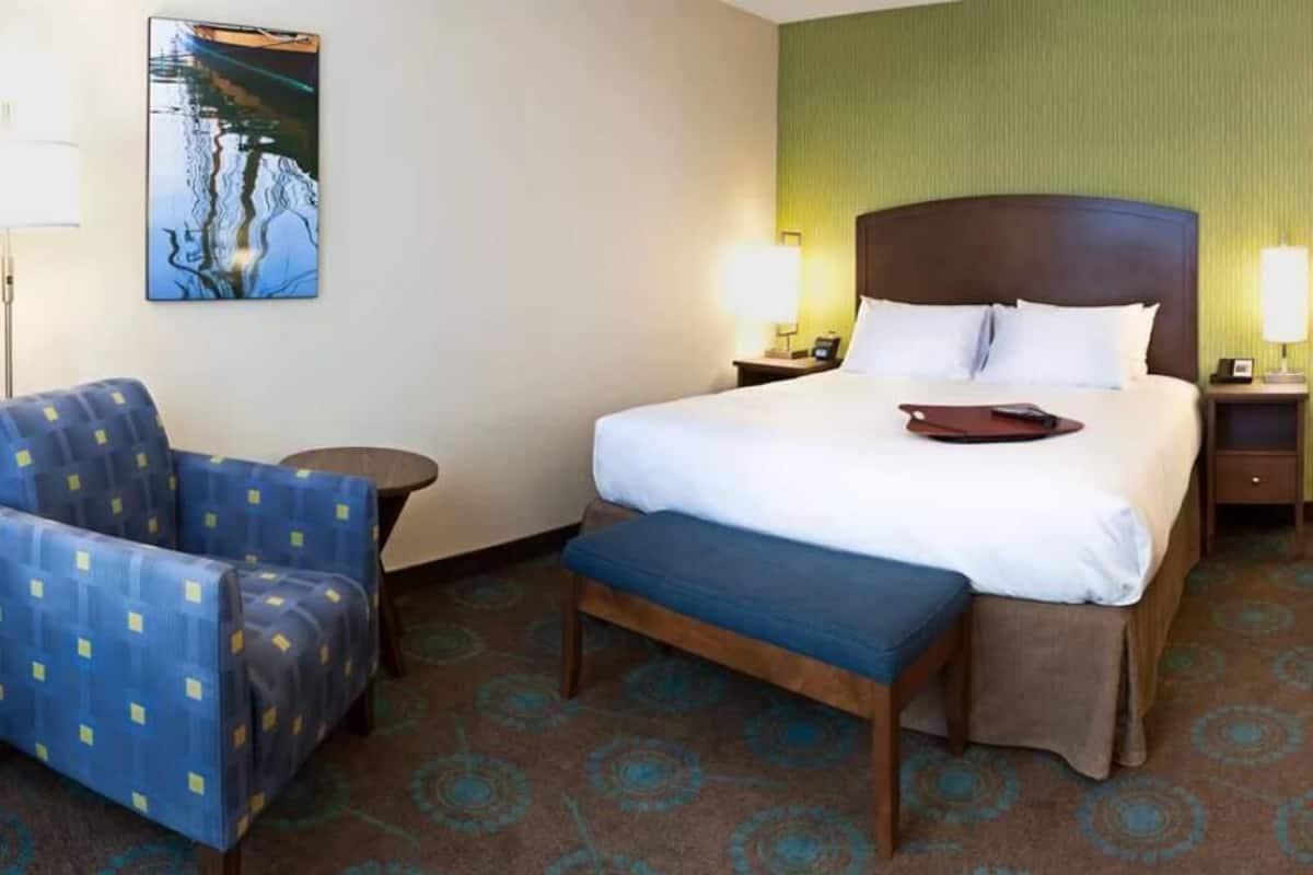 A hotel room with a bed, a chair, a bedside table, a lamp, and a wall-mounted artwork.