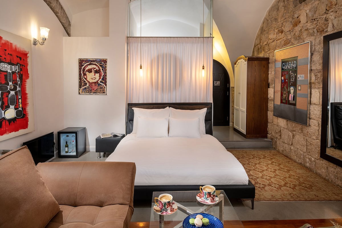 Habitación de hotel con una cama grande, un sofá, una mesa de centro con tazas y un cuenco de macarons, y una pared de piedra.