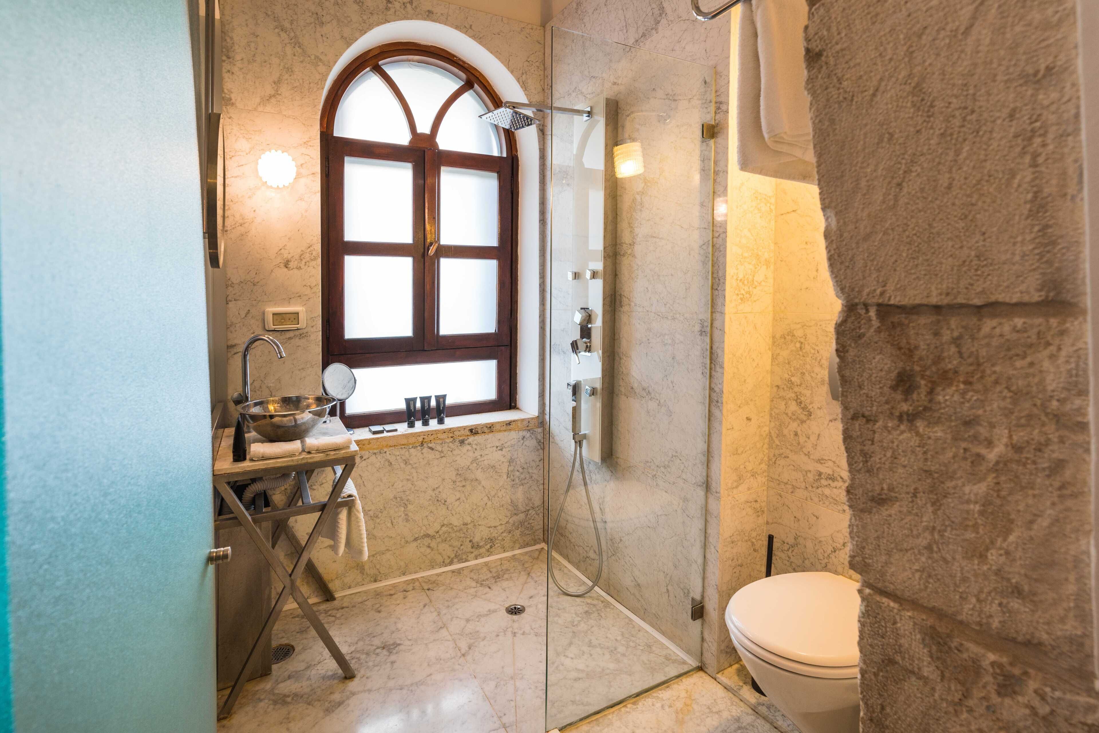 A bathroom with a glass-enclosed shower, a marble floor, a sink with a basin, and a window with a wooden frame.