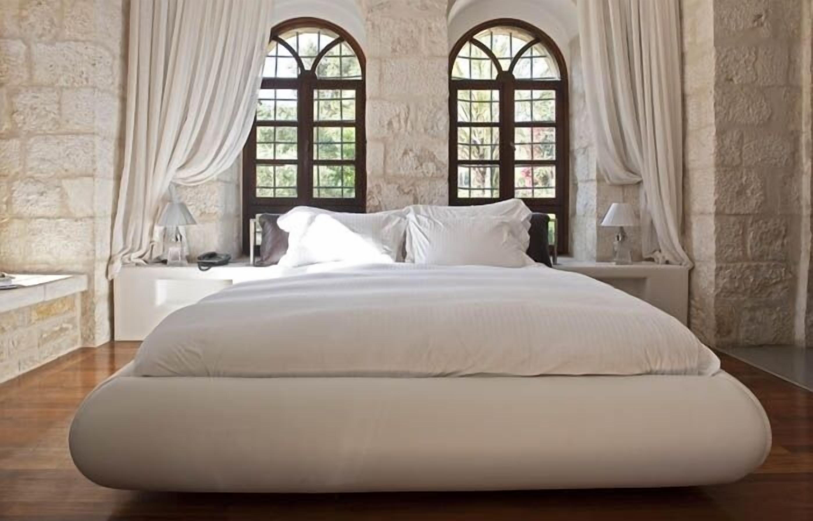 A large bed in a room with stone walls and two arched windows.