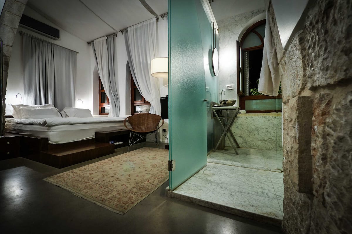 Un dormitorio con cama, una silla, una mesita y un baño visible a través de una puerta abierta.