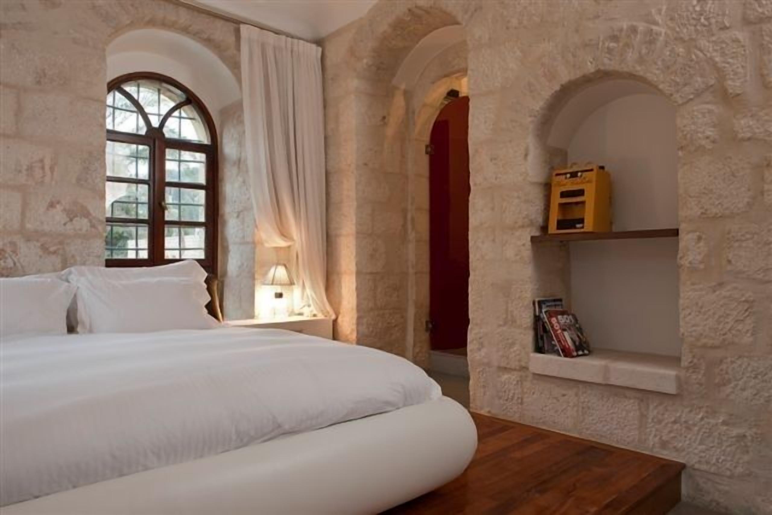 A bedroom with a large bed, a wooden nightstand, a window with curtains, and a stone wall with a shelf.